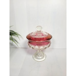 May include: A pedestal candy dish made of clear and cranberry-coloured glass. The bowl and lid feature a pressed pattern of raised dots and geometric shapes. The lid is topped with a small clear glass knob, and the base is a circular pedestal.