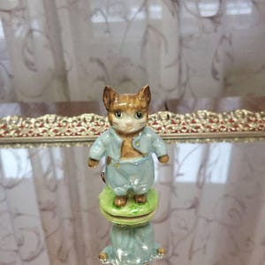 May include: A ceramic cat figurine, wearing a light blue outfit. The cat has brown and tan fur, green eyes, and stands on a green base. The figurine is reflected in a mirrored surface.