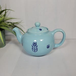 May include: A light blue ceramic teapot with a matching lid. The teapot features a rounded body, a curved spout, and a large handle. Dark blue floral designs are painted on the teapot. The teapot is sitting on a white surface.