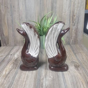 May include: Two brown ceramic swan figurines with white and textured accents. The swans are displayed side-by-side, showcasing their elegant, curved necks and detailed wing designs. The figurines are on a wooden surface.