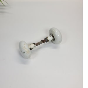 May include: A vintage door knob set featuring two round, white porcelain knobs connected by a central metal spindle. The white finish on the knobs shows signs of wear and ageing, with visible chips and discolouration on the metal components.