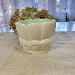 May include: A cream-colored ceramic planter with a scalloped rim and a light green interior. The base features a decorative floral design. The planter is suitable for small plants and adds a touch of vintage charm to any space.