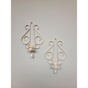 May include: A pair of off-white metal wall sconces featuring a symmetrical scrollwork design. Each sconce has a central vertical frame with curved decorative elements and a small circular platform at the base intended to hold a single candle.