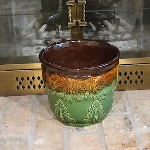 May include: A ceramic planter with a dark brown interior and rim, transitioning to a band of yellow and orange floral relief. The lower portion is green with leaf designs. The planter sits on a brick surface.