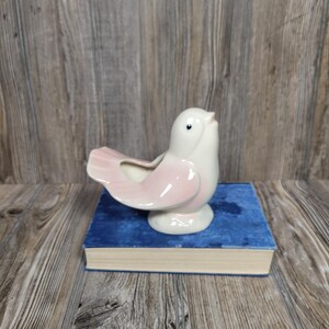 May include: A ceramic bird-shaped planter in white and pink, perched on a blue book. The bird has a detailed design with a small eye and a curved tail. The planter is likely for small plants or flowers.