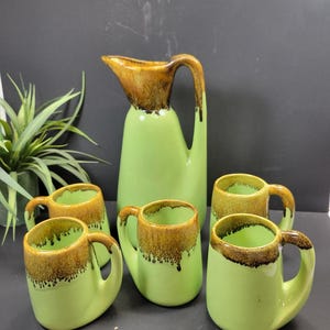 May include: A vintage green ceramic pitcher and matching mugs set. The pitcher has a brown top and handle, while the mugs have brown rims. The set is a retro style, perfect for serving beverages. The mugs are small and have a curved handle.
