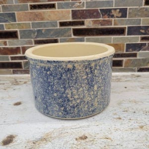 May include: A cylindrical ceramic crock featuring a speckled blue and cream sponge-painted finish. The vessel has a smooth, unglazed cream-colored interior rim and a flat base, presenting a sturdy, open-top design for storage or display purposes.