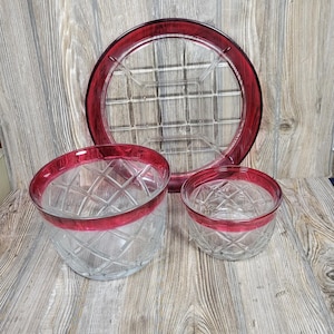 May include: A clear glass serving set with a red rim. The set includes a large round tray and two bowls. The glass has a diamond pattern. The tray has a grid pattern.