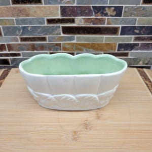 Peut inclure: Un cache-pot ovale en céramique avec un bord festonné et un intérieur vert pâle. L'extérieur est blanc avec un motif de feuilles en relief entourant la base. Le récipient présente une finition lisse et brillante et une ouverture large.