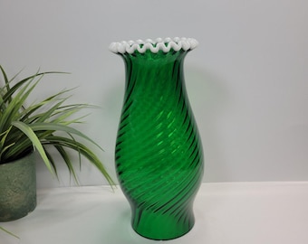 Pode incluir: Um vaso de vidro verde com um padrão canelado em espiral por todo o corpo. O vaso apresenta um pescoço alargado e uma borda de vidro branco com babados na abertura superior. O vidro é translúcido e de cor verde profunda, exibindo uma textura em espiral consistente da base ao topo.