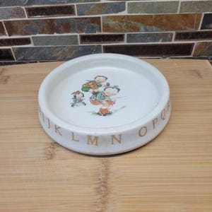May include: A round white ceramic dish featuring a central illustration of three children playing together. The outer rim of the dish is decorated with a series of letters from the alphabet printed in a light brown colour, including I, J, K, L, M, N, O, P, Q.