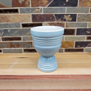 May include: A light blue ceramic egg cup featuring a rounded bowl with horizontal ribbed detailing around the upper section and a matching ribbed pedestal base. The interior of the cup is white, and the exterior has a smooth, glossy finish.