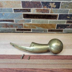 May include: A brass door handle with a spherical end and a curved, tapered body featuring two small pre-drilled holes for mounting. Two small metal screws are positioned directly below the handle on the surface.