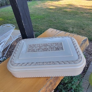 May include: A rectangular, cream-colored storage box with a smooth, glossy finish. The lid features a central rectangular panel and a surrounding border, both decorated with an intricate, embossed floral pattern in a contrasting tan color.