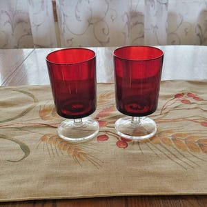May include: Two matching drinking glasses featuring deep red bowls and clear glass stems with circular bases. The glasses have a cylindrical shape with a smooth finish and are designed for serving beverages, showcasing a vibrant ruby color.