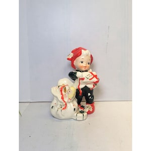 May include: A vintage Christmas figurine of a child elf holding a gift and standing next to a sack of presents. The elf wears a red hat, black pants, and a white shirt. The sack is white with gold accents and red ribbon.