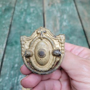 May include: A gold-toned metal drawer pull featuring an oval center plate with a decorative border and a hinged, semi-circular drop handle. The hardware has a weathered, textured finish with dark patina accents in the recessed areas.