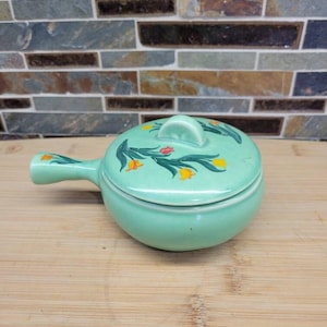 May include: A light green ceramic pot with a matching lid and a single side handle. The exterior of the lid and handle features a painted floral pattern consisting of green stems, dark green leaves, and small red and yellow tulip-like flowers.