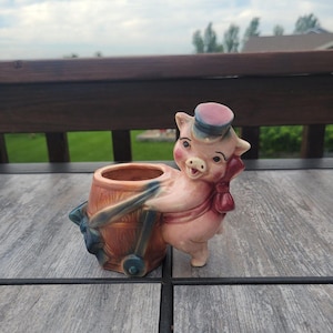 May include: A ceramic figurine of a pink pig wearing a small blue hat and a red neckerchief, pushing a brown wooden-style barrel planter. The pig has painted facial features, and the barrel has a blue wheel and handle accents on the side.