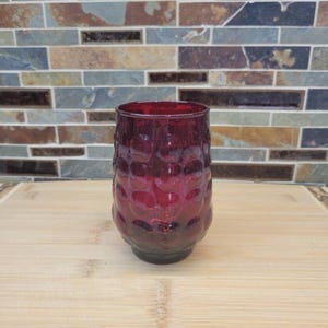 May include: A ruby red glass with a textured, bubble-like pattern. The glass has a rounded shape and a dark base, sitting on a wooden surface. The background features a mosaic tile design.