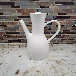 May include: A white ceramic coffee pot with a long spout and a curved handle. The pot has vertical ridges and a silver-colored lid. The background features a multi-colored brick pattern.