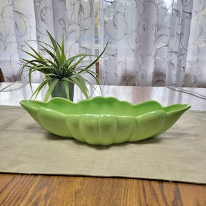 May include: A bright green ceramic serving dish with a scalloped edge, resembling a pea pod. The dish is placed on a beige linen tablecloth, with a small potted plant in the background. The table is made of wood.