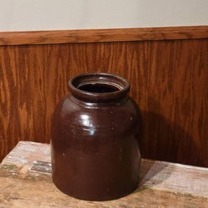 May include: A dark brown ceramic crock with a cylindrical body and a thick, rounded rim. The surface features a glossy, glazed finish with subtle variations in color and texture throughout the exterior of the vessel.