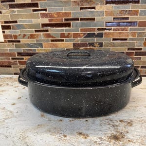May include: An oval-shaped black roasting pan with a speckled white enamel finish. The roaster features a matching lid with a central handle and two side handles on the main body, providing a classic look for cooking large meals in the kitchen.