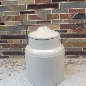May include: A cylindrical off-white ceramic crock with a matching lid and a thin, dark metal wire handle. The container features a smooth, glossy finish and a slightly tapered neck leading to a rounded lid with a small central knob.