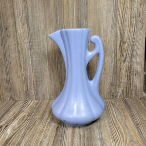 May include: A light periwinkle blue ceramic pitcher featuring a vertical ribbed texture, a flared spout, and a curved handle. The vessel has a wide, rounded base that tapers upward toward the rim, showcasing a smooth, matte finish throughout.