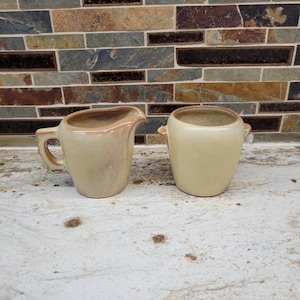 May include: Two vintage ceramic creamers. The creamer on the left has a handle and a spout, with a light tan body and a darker rim. The creamer on the right is a small, round, handled bowl in a similar color. Both are on a white surface.