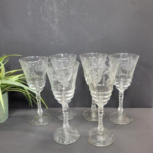 May include: A set of six clear glass goblets featuring etched floral patterns on the bowls. Each goblet has a multi-tiered, ribbed base leading down to a slender, faceted stem and a circular foot. The glassware is transparent and colorless.