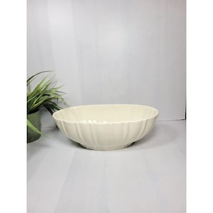 May include: A cream-colored, oval-shaped ceramic bowl with a fluted design. The bowl is set against a white background with a hint of a green plant in a pot on the left side. The bowl's dimensions are not visible.