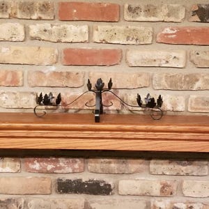 May include: A black metal candle holder with a leaf design. The candle holder is mounted on a wooden shelf in front of a brick wall. The candle holder has three places for candles.