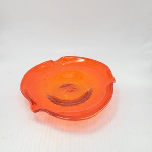 May include: An orange glass ashtray with a textured surface. The ashtray has a circular shape with three curved indentations on the rim. The ashtray is made of a translucent orange glass.