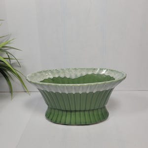 May include: An oval-shaped ceramic planter featuring a deep green base with vertical ribbed detailing. The upper rim is flared and finished with a contrasting white glaze that drips down over the green, creating a scalloped edge effect.