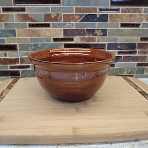 May include: A brown ceramic mixing bowl with horizontal ridges, set on a light-coloured wooden chopping board. The background features a multi-coloured tile backsplash.