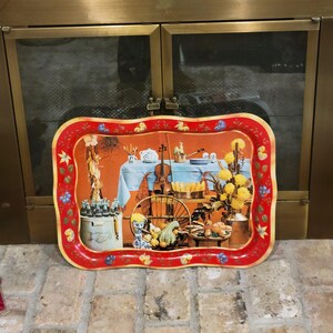 May include: A vintage metal serving tray with a red border and a colorful illustration. The illustration depicts a still life scene with a violin, flowers, and various food items. The tray has a scalloped edge and is set against a brick background.