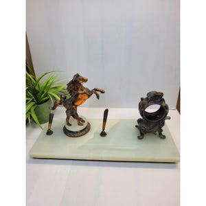 May include: A rectangular light green marble desk set featuring a bronze-toned rearing horse sculpture on a circular base, two slender pen holders, and a separate ornate circular bronze-toned inkwell or pen stand with intricate decorative detailing.