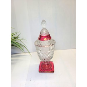 May include: A clear glass jar with a diamond pattern, a red band, and a matching red square base. The jar has a decorative lid with a crystal finial. The jar is likely used for storage or display.