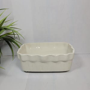 May include: A rectangular off-white ceramic baking dish featuring a scalloped top rim and vertical ribbed detailing along the exterior sides. The vessel has a smooth, glossy finish and a deep, open interior basin for holding various food items.
