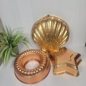 May include: Three vintage copper-colored baking molds: a ring-shaped mold, a star-shaped mold, and a large shell-shaped mold. The molds are made of metal and have detailed designs. The shell mold is approximately 10 inches in diameter.