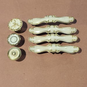 May include: A collection of seven cabinet hardware pieces consisting of three circular knobs and four elongated drawer pulls. Each piece features an off-white finish with gold-toned metallic detailing along the edges and decorative scrollwork.