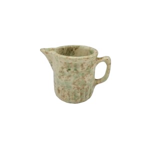 May include: A small ceramic creamer pitcher featuring a speckled pattern in shades of light green and tan. The pitcher has a rounded body, a short pouring spout, and a curved handle on the side. The surface has a textured appearance with visible color variations throughout the glaze.