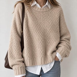 May include: A beige long-sleeve knit sweater with a crew neckline and ribbed cuffs, layered over a white collared button-down shirt and blue denim jeans, accompanied by a dark brown leather shoulder bag with a gold-tone buckle.