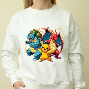 May include: White sweatshirt featuring a colorful graphic of three cartoon characters: a blue and green creature, a yellow character, and a red dragon-like creature. The graphic has a cartoon style with yellow and orange accents.