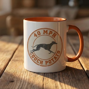 Puede incluir: Taza de cerámica blanca con interior y asa naranja. La taza presenta un diseño circular con una silueta de galgo corriendo, el texto "40 MPH" arriba y "COUCH POTATO" abajo.