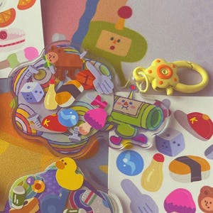 May include: A selection of colourful acrylic charms and sticker sheets. The charms feature various designs, including food items, and are attached to keychains. The sticker sheets showcase illustrations of food, objects, and other playful elements. The background is a vibrant, patterned design.