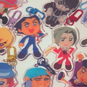 May include: A collection of clear acrylic keychains featuring various cartoon characters with distinct outfits and hairstyles, each attached to a colourful metal lobster clasp. The characters include figures in suits, hats, and casual attire, with one keychain displaying the text Ralumairy.
