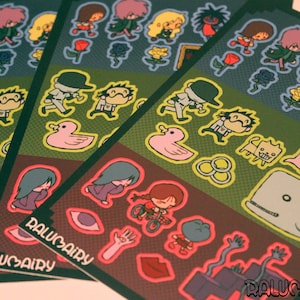 May include: A sticker sheet featuring various cartoon illustrations including characters, a pink duck, a whale, roses, and eyes. The sheet has a dark background with a grid pattern and the text RALUMAIRY printed in a stylised font at the bottom corners.